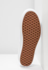 Semelle de chaussure avec motif gaufré en caoutchouc marron et semelle intermédiaire blanche, positionnée verticalement contre un mur et un sol blancs.