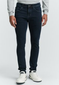 Dunkelblaue Skinny-Jeans mit glatter Textur, Fünf-Taschen-Design und einem dezenten, ausgebleichten Look. Kombiniert mit weißen Sneakers.
