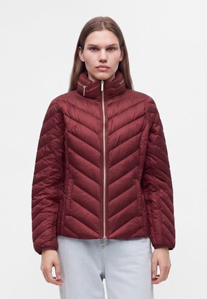 Femme portant une veste matelassée grenat avec un col montant et une fermeture éclair devant, debout devant un fond clair uni.