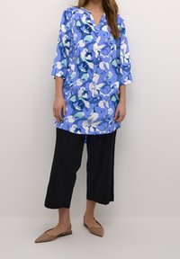 Blumen-Tunika in Blau und Weiß, 3/4-Ärmel, V-Ausschnitt, lockere Passform. Kombiniert mit schwarzen Weitbein-Hosen und beigen Slingback-Schuhen.