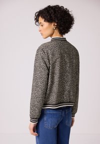 Donna con capelli ricci in jeans blu che indossa una giacca bomber grigia e nera con stampa animalier, polsini e colletto a righe, rivolta verso sinistra.