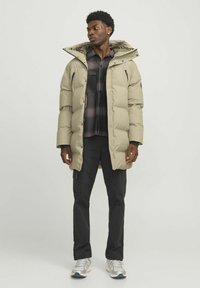 Beige puffer jacket with a zip and snap front, large hood, side pockets, worn over a dark plaid shirt and black cargo pants.