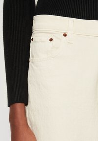 Jeans en coton de couleur crème avec une finition texturée, présentant des fermetures en cuivre et des poches avant. Haut à manches longues en tissu côtelé noir exposé.
