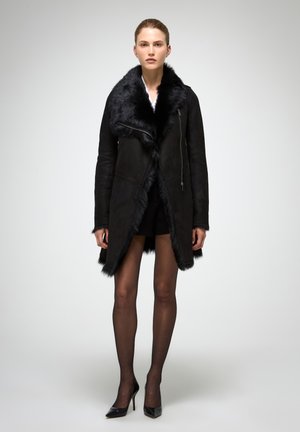 Zwarte suède jas met een grote faux fur kraag en asymmetrische zoom. Voorkant met ritsdetail en lange mouwen. Draagt over een zwarte outfit.