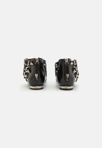 Bottines en cuir verni noir avec une fermeture éclair, présentant des accents métalliques et un talon texturé. La vue arrière montre des détails entrecroisés.