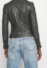 Persoon in een aansluitende zwarte leren jas met ritsen op de mouwen en lichtblauwe high-waisted jeans, weergegeven van achteren.
