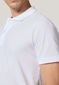 Biała koszulka polo z teksturowanej tkaniny, z krótkimi rękawami i zapięciem na trzy guziki. Posiada klasyczny kołnierz oraz dopasowany krój.