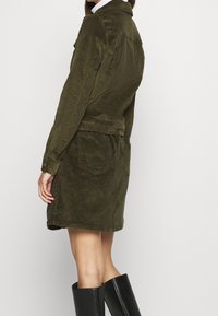 Woman wearing olive green corduroy jacket and matching skirt with back pockets, paired with black knee-high boots, against white background.