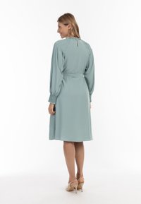 Rochie verde mentă cu mâneci lungi, detalii plisate la umeri și încheieturi, panglică la talie și tiv sub genunchi. Poartă cu pantofi cu toc nude.
