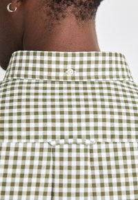 Il colletto della camicia a scacchi verdi e bianchi presenta una chiusura con bottone sul retro. Il tessuto appare liscio con una vestibilità strutturata.