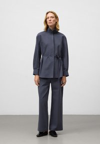 Kvinna som bär en mörkgrå, bälte-häftad högkragejacka med matchande vida byxor och svarta slip-on skor, stående mot en enfärgad bakgrund.