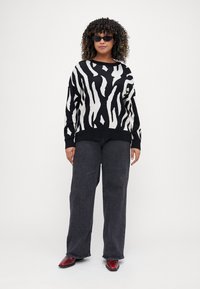 Μαύρος και λευκός ζεβρέ πουλόβερ με χαλαρή εφαρμογή, συνδυασμένο με σκούρα τζιν και κόκκινα παπούτσια. Το μοντέλο φορά μικρά μαύρα γυαλιά ηλίου.