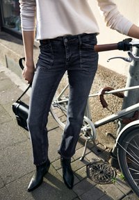 Černé džíny z denimu s mírným vyblednutím, s předními kapsami a přiléhavým střihem. Kombinovány s černými špičatými kotníkovými botami a držící tašku.