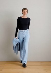 Μαύρη ριγέ μπλούζα μακρύ μανίκι, ανοιχτό μπλε παντελόνι ψηλής μέσης και σκούρα παπούτσια. Ένα τζην μπουφάν κρατιέται στο ένα χέρι. Το έδαφος είναι ξύλινο.