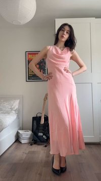 Γυναίκα που φοράει ένα ροζ σατέν φόρεμα χωρίς μανίκια και μαύρα ψηλοτάκουνα στέκεται με τα χέρια στη μέση σε μια κρεβατοκάμαρα με κιθάρα και κορνιζαρισμένη αφίσα.