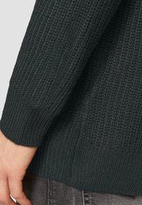 Dunkelgrüner Strickpullover mit strukturiertem Rippenmuster und breiten Bündchen, auf dem ein Teil einer Hand liegt, die auf einem Denim-Stoff ruht.