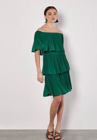 Grünes, plissiertes, schulterfreies Kleid mit gestuften Rüschen, verfügt über eine taillierte Taille und einen ausgestellten Saum. Kombiniert mit braunen Sandalen.