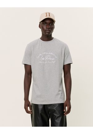Camiseta de algodón gris con mangas cortas, con texto en blanco en el centro del pecho. El modelo también lleva una gorra beige con un logo.