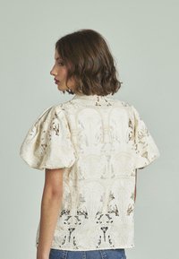 Spitzenbluse in Elfenbein mit puffärmeligen kurzen Ärmeln und kunstvollen floralen Mustern. Mit Rundhalsausschnitt und transparenter Textur, kombiniert mit Denim.