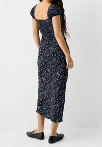 Svart blommig midiklänning med blåa detaljer, fyrkantig halsringning, korta puffärmar, figursydd kropp och svängd kjol. Buren med svarta platta skor.