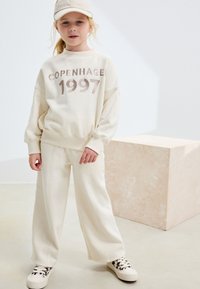 Sweat-shirt crème avec l'inscription « COPENHAGEN 1997 » en lettres brunes, assorti à un pantalon large coordonné et des baskets claires avec un accent imprimé léopard.