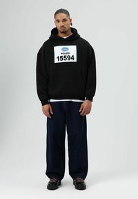 Zwarte hoodie met grote witte opdruk "POLIZEI 15594" op de voorkant, gecombineerd met losse donkere spijkerbroek en zwarte schoenen met roze veters.