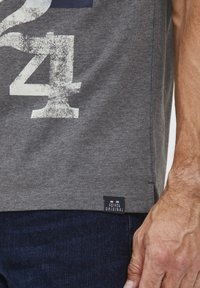 Grå bomulls-t-skjorte med et stort, falmet hvitt tall "24" trykk, med et merke-tag nederst til venstre. Båret med mørk denimjeans.