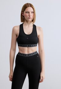 Μαύρο αθλητικό crop top με στρογγυλή λαιμόκοψη, ραβδωτή υφή και λευκή κορδέλα με λογότυπο. Συνδυάζεται με αντίστοιχο κολάν ψηλής μέσης.