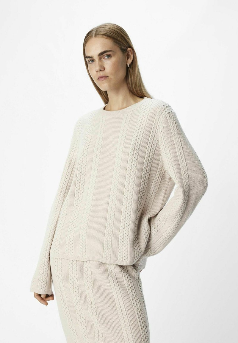 Maglione a maglia beige chiaro con motivi a cavo verticali, collo a giro e maniche lunghe, abbinato a pantaloni in maglia testurizzata coordinati.