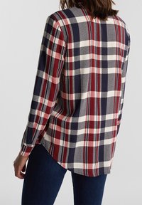 Donna che indossa una camicia a maniche lunghe a scacchi rossi, blu navy e bianchi, con jeans blu scuro, vista da dietro su uno sfondo semplice.