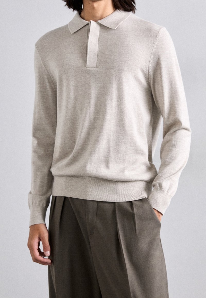 Pull polo beige à manches longues avec une patte de boutonnage sur le devant, des poignets côtelés et une coupe décontractée. Fabriqué dans un tissu doux et lisse. Associé à un pantalon gris.