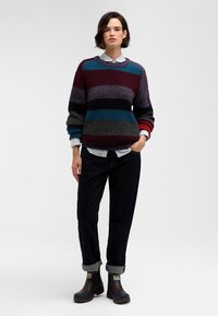 Multicolored knit sweater with horizontal stripes in maroon, teal, gray, and black, layered over a collared striped shirt, paired with dark pants and brown boots.