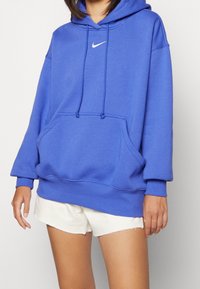 Blå hoodie i mjukt tyg, med framficka, dragsko i huvan och en liten vit Nike-logga på bröstet.