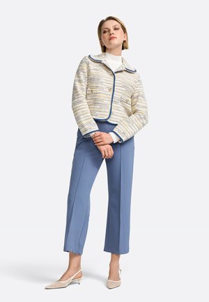 Veste en crème texturée avec des accents bleu clair, à double boutonnage à l'avant, associée à un pantalon bleu sur mesure et des chaussures à talons ouverts beiges.