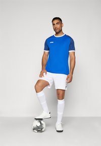 Maglia azzurra a maniche corte con colletto bianco e maniche con fantasia, abbinata a pantaloni corti bianchi e scarpe da calcio nere, in piedi su una palla da calcio.