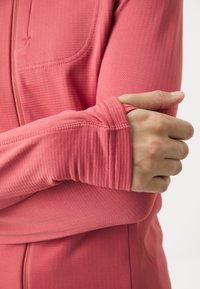 Acercamiento de una persona que lleva una camiseta de manga larga color coral con agujero para el pulgar, con el brazo cruzado y la mano apoyada en el brazo contrario.