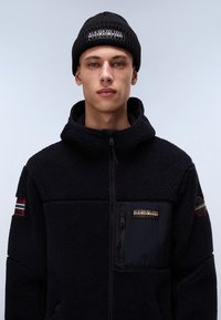Black fleece jacket with a zipper, high collar, and logo details; features a textured fabric and a black knit beanie with branding.