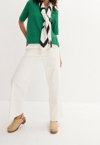 Top corto a maniche corte in maglia verde con motivi a occhiello, abbinato a pantaloni bianchi a gamba larga e zoccoli in legno color nocciola con accenti metallici.