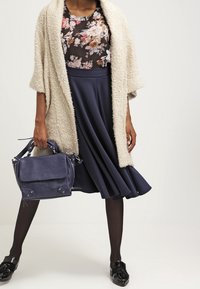 Cardigan beige con texture sopra una maglietta trasparente floreale, abbinato a una gonna a campana blu scuro. Tiene in mano una borsa in pelle blu scuro e indossa scarpe nere.