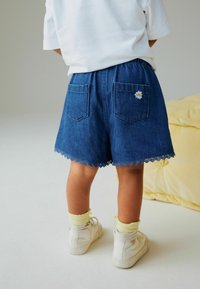 Spijkershorts met een blauwe tint, geplooide zoom, geborduurde madelief op achterzak, elastische tailleband, gecombineerd met witte schoenen en gele sokken.