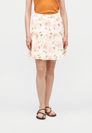 Žena v bielej kvetinovej sukni, oranžovom tielku bez rukávov, zlatých náramkoch a hnedých sandáloch s remienkami, stojaca na jednolennom pozadí.