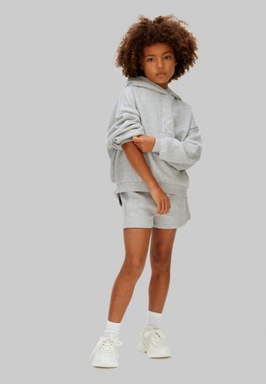 Kind mit lockigem Haar, trägt einen grauen Kapuzenpullover und passende Shorts, steht mit einem Arm über dem Körper, weiße Socken und Turnschuhe.