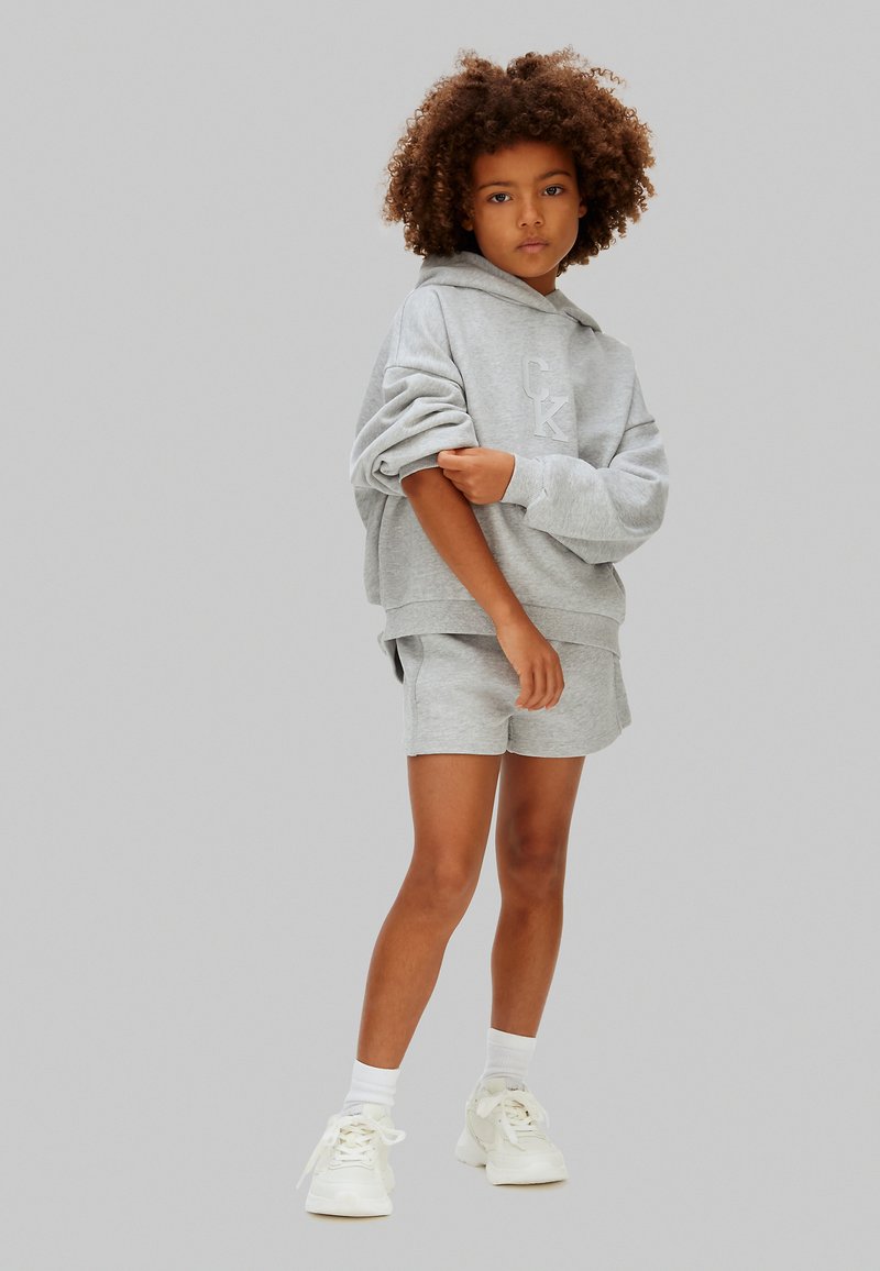 Enfant aux cheveux bouclés portant un sweat à capuche gris et un short assorti, debout avec un bras croisé sur le corps, chaussettes blanches et baskets.