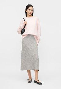 Žena v svetloružovom svetri, šedej midi sukni s vrkôčikmi, čiernych balerínach a s čiernou kabelkou cez rameno, stojaca pred jednoduchým pozadím.