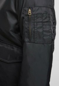 Schwarze Bomberjacke mit einer Reißverschlusstasche am linken Ärmel, strukturiertem Stoff und auffälligen Nähdetails.