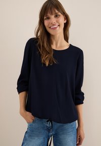 Haut à manches longues bleu marine avec un col rond, présentant une texture côtelée subtile et un détail froncé au niveau de l'ourlet, associé à un pantalon en denim clair.