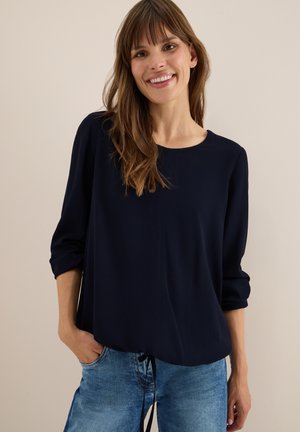 Navyblauwe longsleeve top met een ronde hals, voorzien van een subtiele ribbelstructuur en een geraffineerd detail aan de onderkant, gecombineerd met lichte denim jeans.