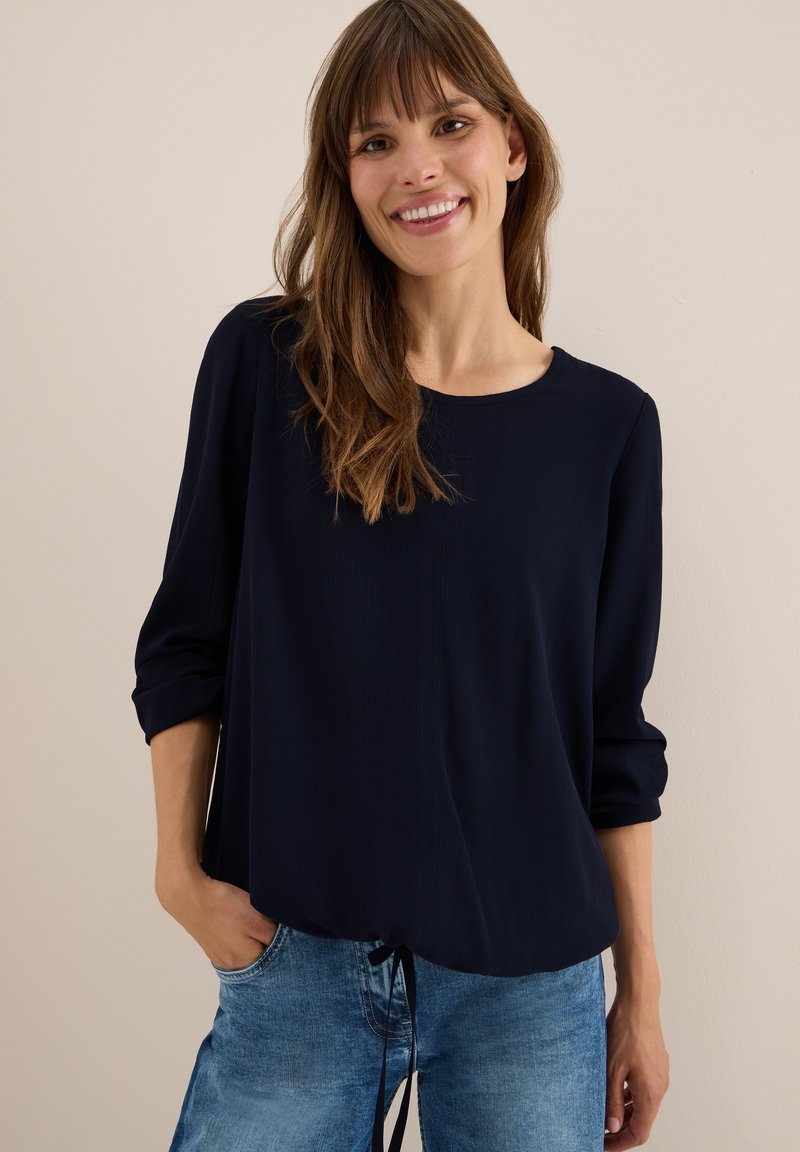 Haut à manches longues bleu marine avec un col rond, présentant une texture côtelée subtile et un détail froncé au niveau de l'ourlet, associé à un pantalon en denim clair.