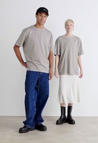 Dva modely nosí světle šedé oversized trička; jeden má na sobě tmavé džíny, druhý krémovou plisovanou sukni a černé robustní boty.