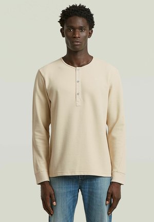 Ung mand stående iført en beige langærmet waffle-strikket henley-shirt og blå jeans mod en ensartet lys baggrund.